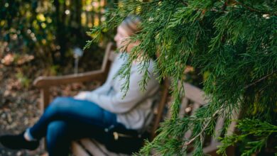 the girl is sitting on a bench near a coniferous tree