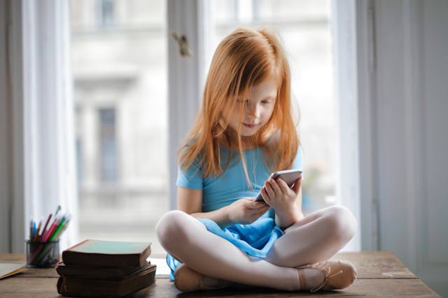 calm-small-ginger-girl-sitting-on-table-and-using-smartphone-in-light-living-room